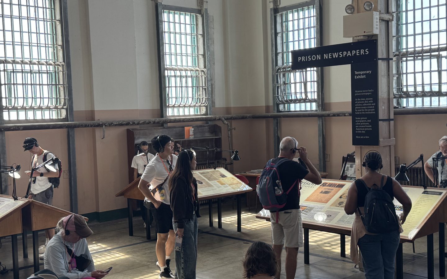 The exhibit installed in the Alcatraz cellhouse: angled reading tables under lamps, visitors standing and sitting at them, and a hanging banner reading “Prison Newspapers — Temporary Exhibit”